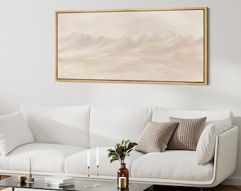 Pode incluir: Pintura de paisagem emoldurada retratando uma cordilheira em tons bege suaves. A obra de arte é exibida acima de um sofá branco com almofadas decorativas. Uma mesa de centro preta com velas e um vaso com vegetação.