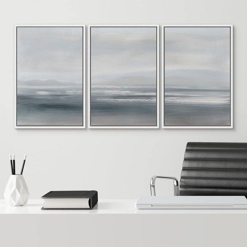 May include: Three framed canvas prints with a white frame. The prints are a light blue and gray color and depict a seascape with waves and a distant shoreline.