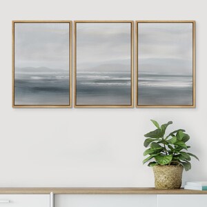May include: Three framed canvas prints with a seascape theme. The prints are in shades of gray and white, depicting a horizon with a distant coastline and waves. The prints are arranged in a row on a white wall above a wooden shelf with a potted plant and books.