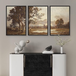 May include: Three framed landscape paintings in a triptych arrangement. The artwork depicts trees, fields, and a body of water in muted tones. Below, a white sculpture, a black bowl, and a vase with white flowers sit on a black and white cabinet.