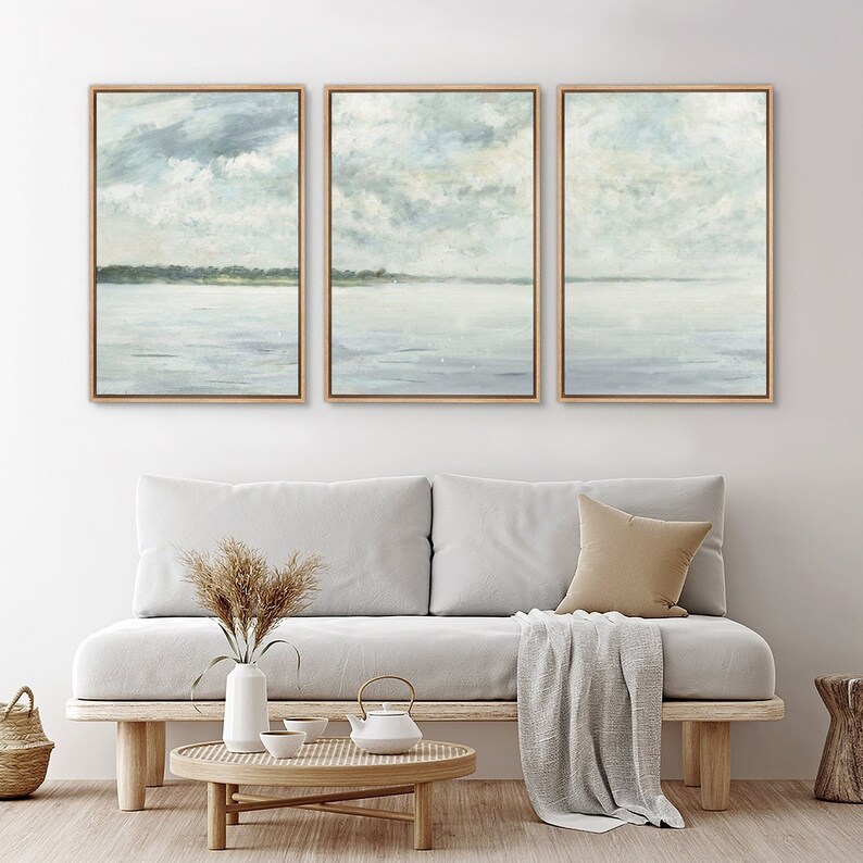 May include: A set of three framed abstract landscape paintings with a muted blue and green color palette. The paintings depict a seascape with a horizon line and clouds in the sky.