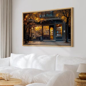 May include: A framed print of a cozy bookstore at night. The shop is lit with warm lights, and the shelves are filled with books. Trees with golden leaves frame the building. The print is displayed above a white sofa.