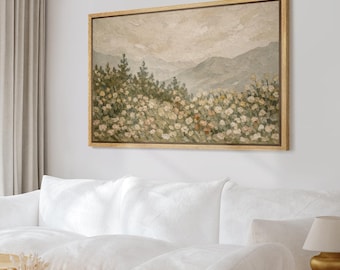 May include: A framed landscape painting featuring a field of flowers in the foreground, with mountains and a cloudy sky in the background. The artwork is in muted tones of beige, green, and gray, with a light brown wooden frame. The painting is displayed above a white sofa.