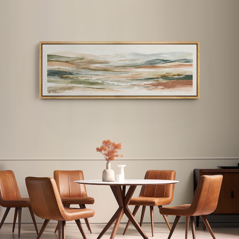 May include: An abstract landscape painting in a gold frame. The artwork features a blend of earthy tones, including greens, browns, and beige, creating a serene and naturalistic scene. The painting hangs above a dining table with brown leather chairs.