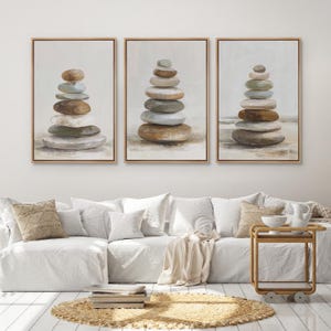 May include: A set of three framed wall art prints, each featuring a vertical stack of smooth, rounded river stones in shades of gray, brown, and beige, set against a neutral, light-colored background with a thin, light-toned wooden frame.
