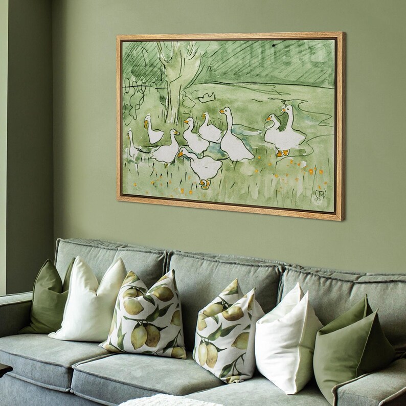 May include: A framed watercolor painting depicts a flock of white geese in a green field. The artwork has a light brown wooden frame and is displayed above a gray sofa with decorative pillows in shades of green and white.