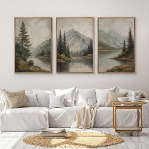 May include: Set of three framed landscape paintings. Each features a serene lake scene with mountains and evergreen trees. The artwork is in muted tones of gray, brown, and green, with a natural wood frame. The paintings are displayed above a white sofa.