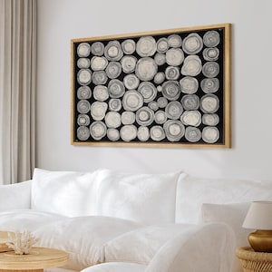 May include: A rectangular wall art piece featuring a black background filled with numerous circular wood slice illustrations in shades of white, gray, and beige, all enclosed within a light-toned wooden frame.