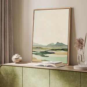May include: A framed landscape painting with a light wood frame. The artwork depicts a serene scene with mountains, water, and fields in shades of green, beige, and blue. An open book rests on a green cabinet.