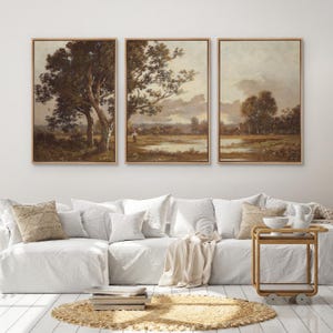 May include: Three framed landscape paintings with a vintage aesthetic. Each features a serene outdoor scene with trees, water, and a muted color palette of browns, greens, and creams. The paintings are displayed above a white sofa with decorative pillows.