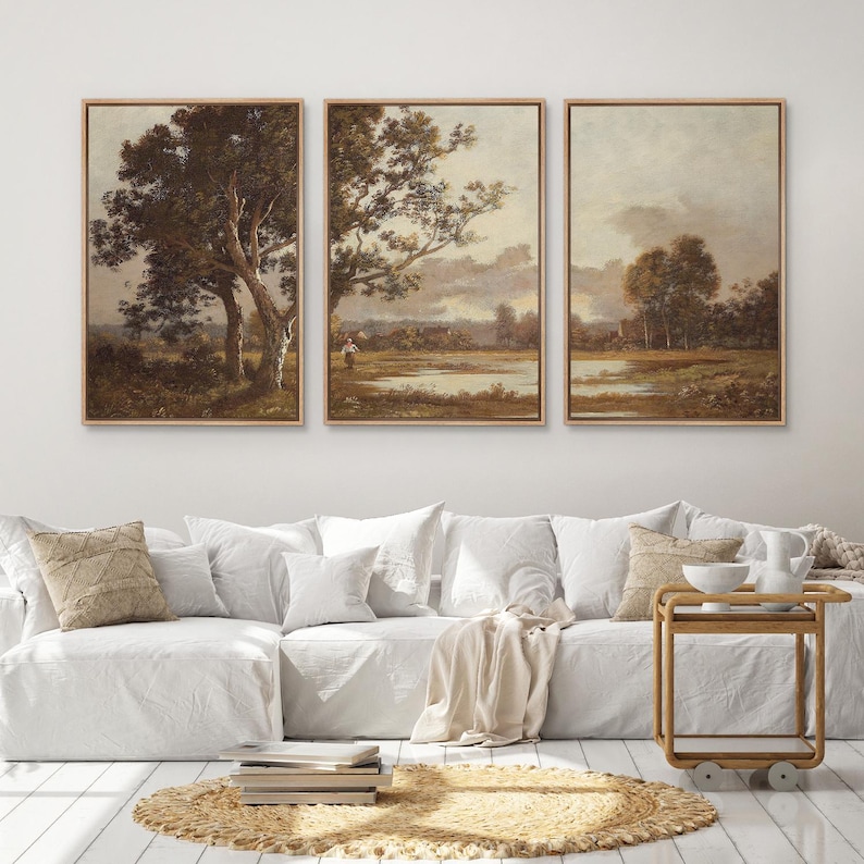 May include: Three framed landscape paintings with a vintage aesthetic. Each features a serene outdoor scene with trees, water, and a muted color palette of browns, greens, and creams. The paintings are displayed above a white sofa with decorative pillows.