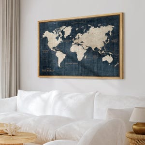 May include: A framed world map print. The map features a vintage design with a navy blue background and beige continents. The frame is made of light brown wood. The text "MAP OF THE WORLD" is visible at the bottom left.