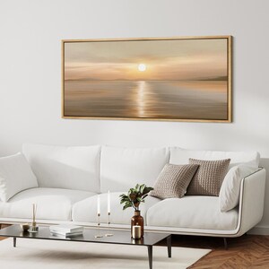 May include: A rectangular landscape painting in a thin gold frame depicting a sunset over a calm body of water. The scene features soft beige, tan, and golden hues with a bright sun centered above the horizon, casting a vertical reflection.