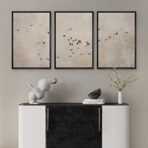 May include: Three framed prints of a flock of birds flying in formation against a light beige background. The prints are arranged in a row on a wall above a white and black cabinet.