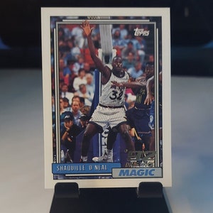 May include: A vintage Topps basketball trading card featuring Shaquille O'Neal in a blue and white Orlando Magic uniform, with the text "Shaquille O'Neal" and "Magic" printed on the card.