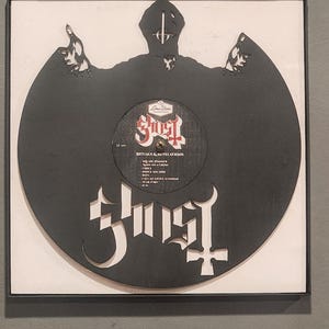 May include: A black vinyl record art piece featuring the band Ghost. The record is cut into the shape of a figure with arms raised. The album title "Rituals & Revelations" is visible on the record label. The artwork is framed in a black frame.