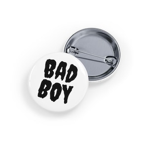 May include: Two white, circular badges with a metal pin fastening. One badge features the words "BAD BOY" in bold, black lettering. The other badge is visible in the background.