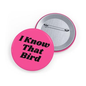 May include: A round pinback button with a bright pink background and the black text I Know That Bird printed in a bold serif font. The reverse side of the button is visible, showing a silver metal backing with a horizontal pin mechanism attached.