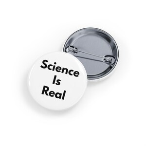 May include: Two white, circular badges with the text "Science Is Real" in black. One badge is face up, and the other is face down, showing the pin back.