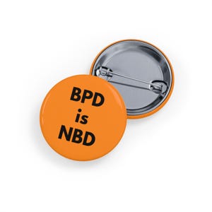 Może przedstawiać: Dwa pomarańczowe, okrągłe guziki z czarnym napisem "BPD is NBD". Jeden guzik jest skierowany w górę, a drugi w dół, pokazując srebrny tył.