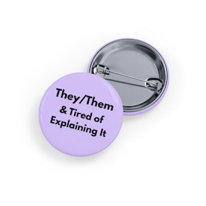 May include: Two round, pale purple badges. One badge has the text "They/Them & Tired of Explaining It" in black. The other badge is silver with a pin back. The badges are on a white background.