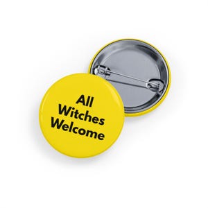 Puede incluir: Dos insignias redondas amarillas con respaldos de alfiler de metal. Una insignia muestra el texto "All Witches Welcome" en negro. La otra insignia es parcialmente visible, mostrando el respaldo de alfiler de metal.