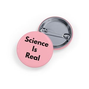 May include: Two round, pink badges with the text "Science Is Real" in black. One badge is face up, and the other is face down, showing the pin back. The badges are likely for a backpack, jacket, or other accessory.