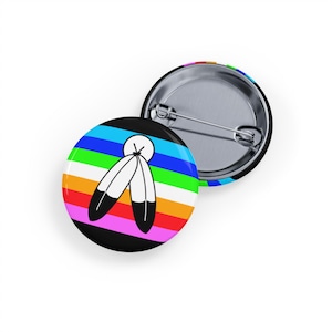 Può includere: Due spille rotonde. Una ha uno sfondo nero con strisce arcobaleno e due piume bianche con punte nere. L'altra mostra il retro della spilla e un design a strisce arcobaleno.