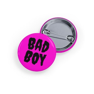 May include: Two round, bright pink pin badges. The front badge has the words "BAD BOY" in bold, black letters. The back of the badge is silver with a pin. One badge is in front of the other.
