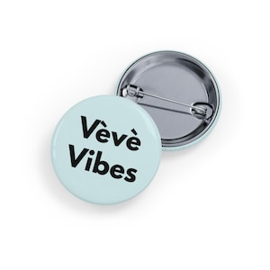May include: Two round pinback buttons. One button is light blue with the words "Vèvè Vibes" in black. The other button is silver. The buttons are likely used as accessories or for decoration.