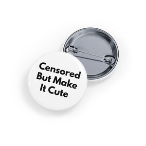 May include: Two white round buttons with a metal pin backing. One button displays the text "Censored But Make It Cute" in black, bold font. The other button is partially visible, showing the metal pin backing.