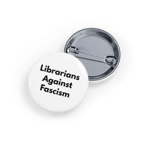 Puede incluir: Dos insignias redondas blancas con cierre de alfiler. Una insignia muestra el texto negro "Librarians Against Fascism". La otra insignia se muestra desde la parte posterior, con un alfiler y cierre plateados.