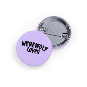 Puede incluir: Dos botones redondos de color lila claro. El botón frontal tiene las palabras "WEREWOLF LOVER" en letras negras y en negrita. La parte posterior de un botón es visible, mostrando un alfiler y respaldo plateados.