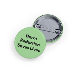 Puede incluir: Dos insignias redondas de color verde claro con el texto "Harm Reduction Saves Lives" en negro. Una insignia está boca arriba y la otra está ligeramente detrás, mostrando el alfiler.
