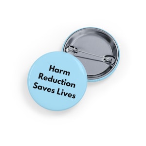 Puede incluir: Dos insignias redondas de color azul claro. Una insignia muestra el texto "Harm Reduction Saves Lives" en negro. La otra insignia es plateada con un alfiler en la parte posterior. Las insignias están sobre un fondo blanco.