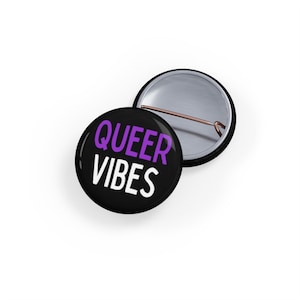 May include: A black button with a white inscription that reads "QUEER VIBES" in purple lettering.
