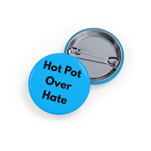 Può includere: Due bottoni rotondi azzurri con la scritta "Hot Pot Over Hate" in nero. Un bottone è rivolto verso l'alto, l'altro verso il basso, mostrando il retro argentato.
