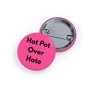 Può includere: Due bottoni rotondi rosa con retro a spillo in metallo. Un bottone mostra il testo "Hot Pot Over Hate" in lettere nere e in grassetto. L'altro bottone è parzialmente visibile, mostrando il retro a spillo in metallo.