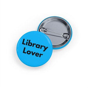 May include: Two round, blue pin badges. One badge is face up, displaying the text "Library Lover" in black. The other badge is face down, showing the silver pin back. The badges are on a white surface.