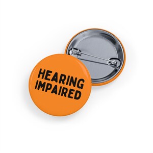 May include: Two orange round badges with the words "HEARING IMPAIRED" in black. One badge is in the foreground, and the other is partially visible in the background. The badges have a silver pin backing.