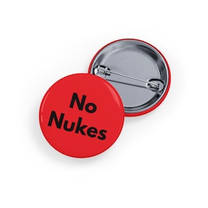 May include: Two red round buttons with the text "No Nukes" in black. One button is face up, displaying the text, and the other is face down, showing the pin back. The buttons are likely made of metal.