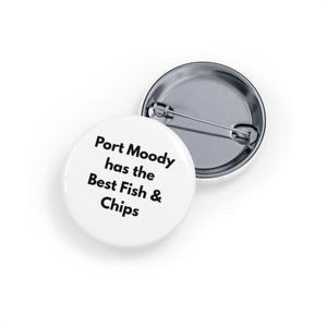 Puede incluir: Dos insignias redondas blancas. Una insignia muestra el texto "Port Moody has the Best Fish & Chips" en negro. La otra insignia es plateada y tiene un alfiler en la parte posterior. Las insignias están sobre una superficie blanca.