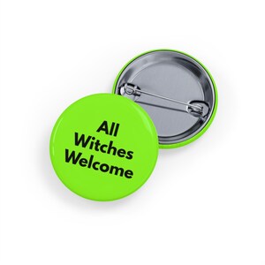 Puede incluir: Dos botones redondos de color verde lima con el texto "All Witches Welcome" en negro. Un botón está boca arriba y el otro boca abajo, revelando un alfiler plateado.