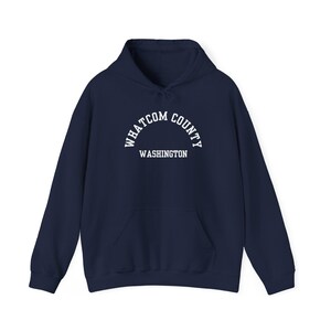 Könnte beinhalten: Marineblauer Kapuzenpullover mit Fronttasche und Kordelzugkapuze. Der weiße Text in Bogenform lautet "WHATCOM COUNTY WASHINGTON". Ein lässiges, bequemes Sweatshirt für den Alltag.
