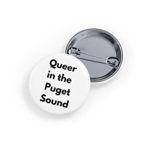 May include: Two white round buttons with a metal pin backing. One button is face up, displaying the text "Queer in the Puget Sound" in black, bold font. The other button is face down, showing the metal pin backing.