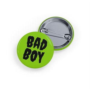 May include: Two round green pin badges. One badge features the words "BAD BOY" in black, bold letters. The other badge is silver with a pin. The badges are on a white background.