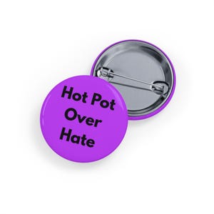 Può includere: Due bottoni rotondi viola con la scritta nera "Hot Pot Over Hate". Un bottone è completamente visibile, mentre l'altro è parzialmente visibile, mostrando il retro della spilla. I bottoni sono su sfondo bianco.