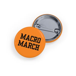 Puede incluir: Dos insignias redondas de color naranja con el texto "MACRO MARCH" en negro. Una insignia está boca arriba, mostrando el texto, y la otra está boca abajo, mostrando el alfiler plateado.