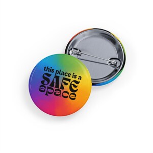 Puede incluir: Un botón de color arcoíris con el texto "this place is a safe space" en negro.