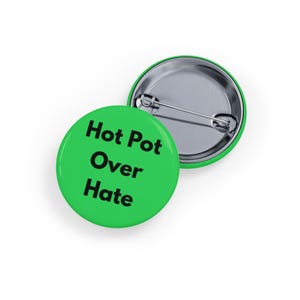 May include: Two green round badges with the text "Hot Pot Over Hate" in black. One badge is face up, and the other is face down, showing the pin back. The badges are on a white background.